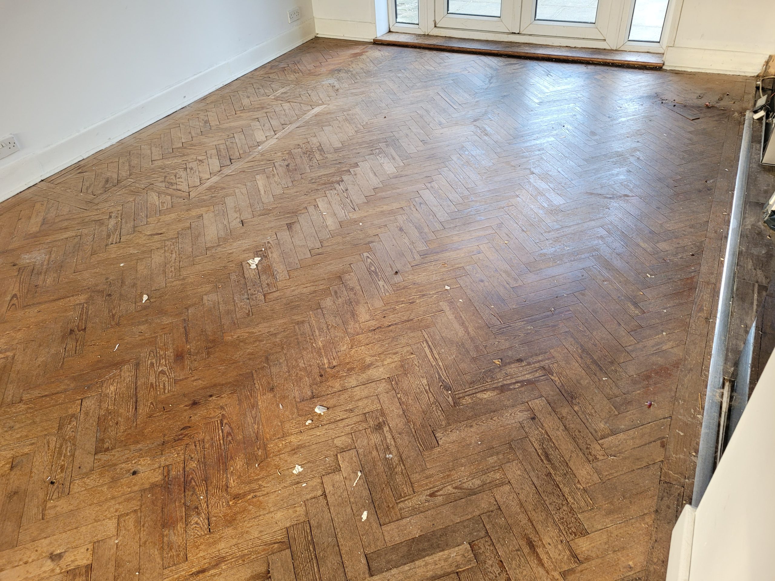 Restoring parquet floors in Harrow – pre-restoration view of a worn herringbone oak parquet living room floor with scratches, gaps and a dull, aged finish.