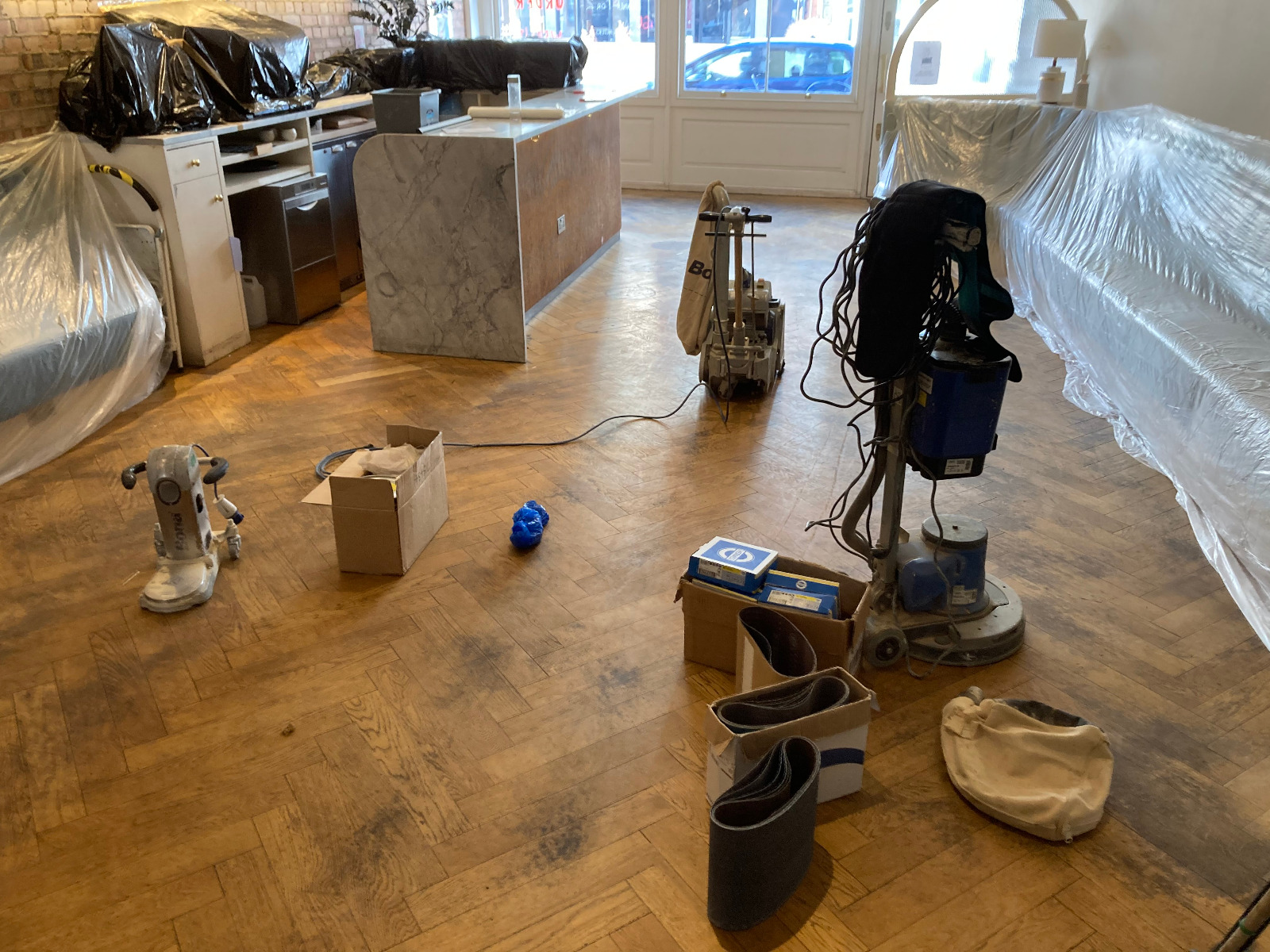Restoring parquet floors in Harrow – preparation scene with dust-sheeted furniture and professional sanding machines ready to work on a herringbone oak parquet wood floor.