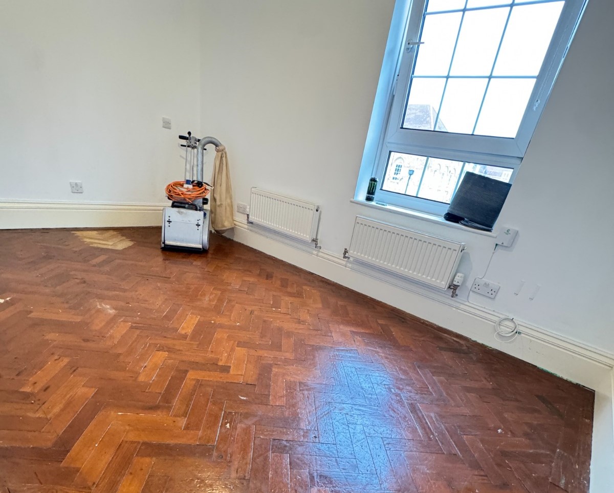 Parquet floor sanding in Harrow on a worn dark oak herringbone parquet wood floor, with a sanding machine set up in an empty room before restoration.